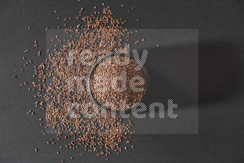 A black pottery bowl full of flaxseeds surrounded by the seeds on a black flooring