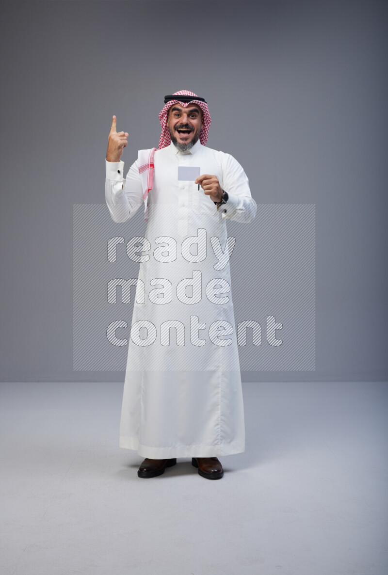 Saudi man Wearing Thob and red Shomag standing holding ATM card on Gray background