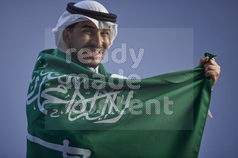 A close-up shot of Saudi man wearing thob and white shomag wrapping big Saudi flag on gray background