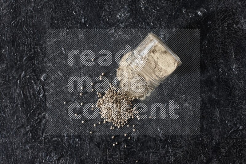 A flipped glass jar full of white pepper powder with spilled powder and pepper beads on textured black flooring