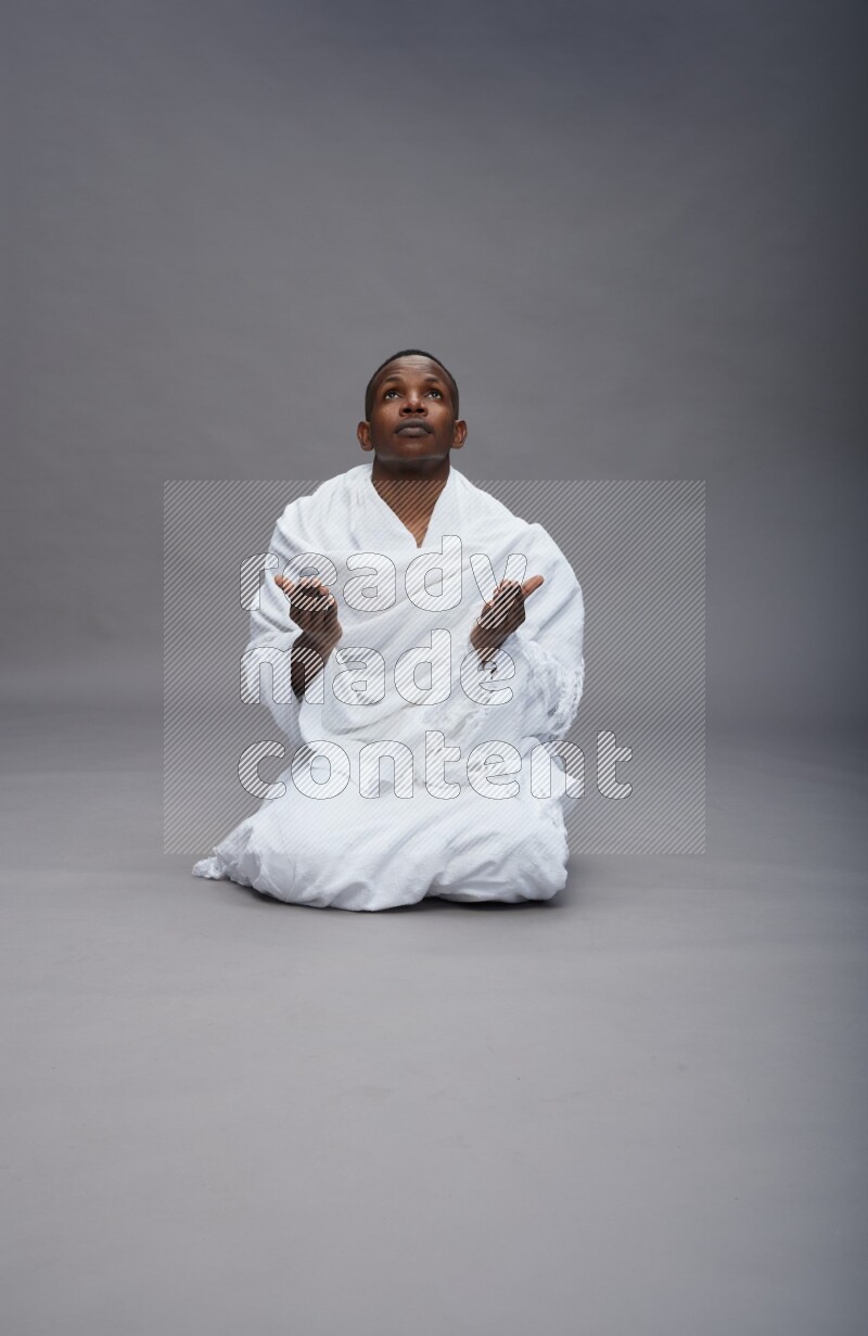 A man wearing Ehram sitting on floor dua'a on gray background