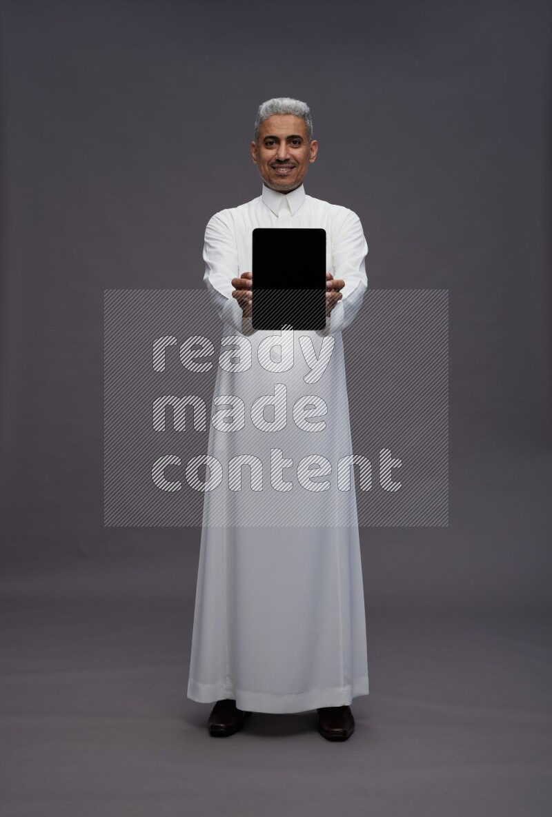 Saudi man wearing thob standing showing tablet to camera on gray background