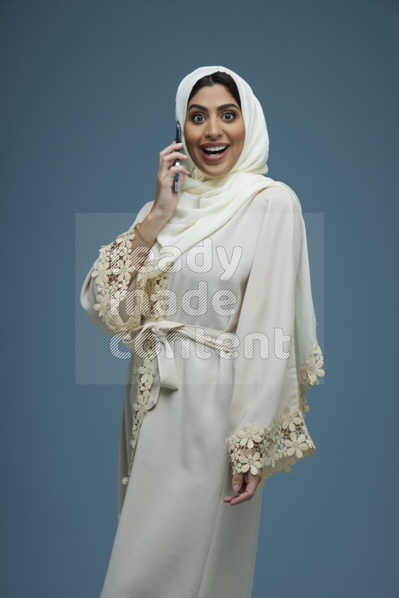 A Saudi woman having a call in a blue background wearing an off-white Abaya Hijab