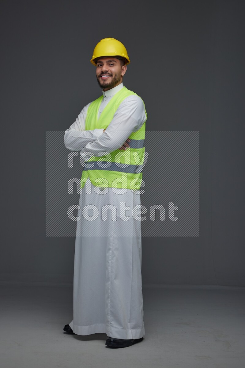 A Saudi man wearing Thobe with a yellow safety vest and white helmet standing and crossing his hands eye level on a gray background