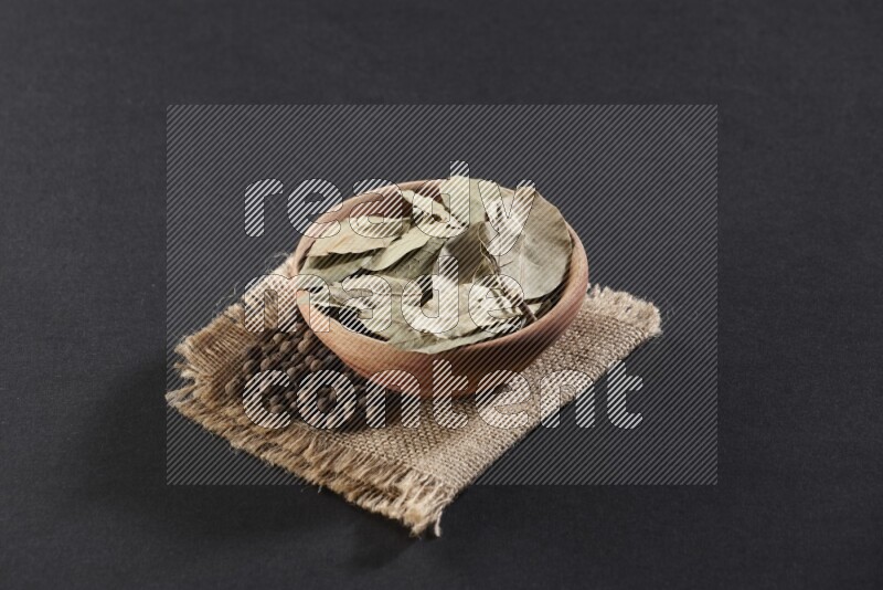 A wooden bowl filled with dried bay leaves on a piece of burlap with bunch of allspice berries on black flooring