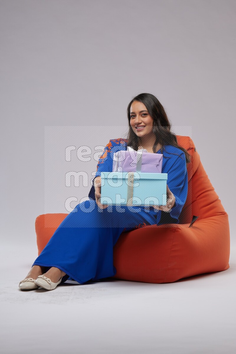 A woman sitting on an orange beanbag wearing Jalabeya holding a gift box