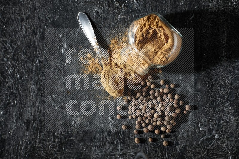 وعاء زجاجي للتوابل مقلوب وملعقة معدنية ممتلئان ببودرة بهار الفلفل الحلو والبودرة تناثرت من الوعاء مع بعض الحبوب على خلفية سوداء