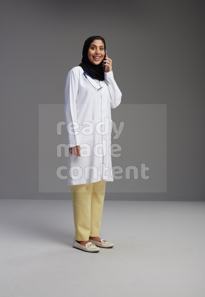 Saudi woman wearing lab coat with stethoscope standing talking on phone on Gray background