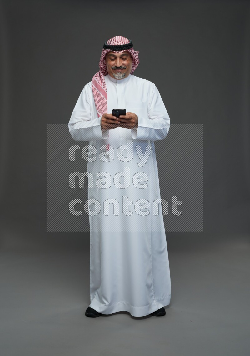 Saudi man with shomag Standing texting on phone on gray background