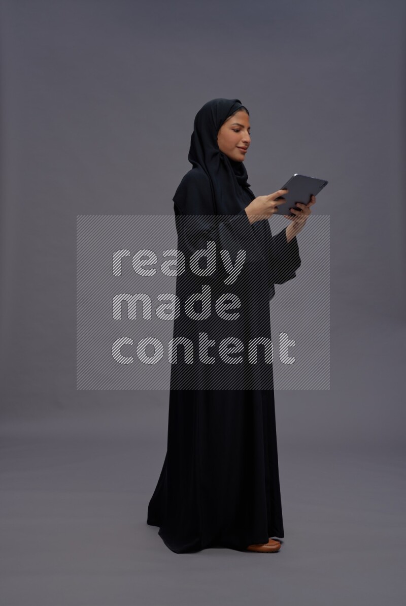 Saudi woman wearing Abaya standing working on tablet on gray background
