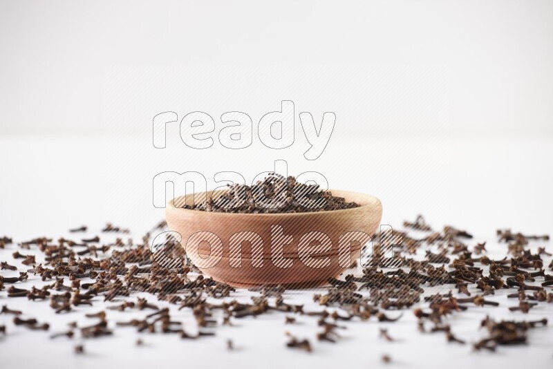 A wooden bowl full of cloves with spread grains on a white flooring