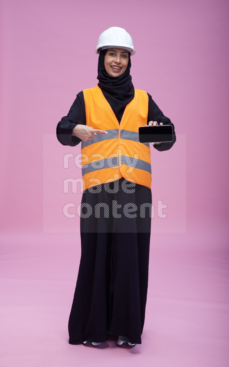 Saudi woman wearing Abaya with engineer vest and helmet standing showing phone to camera on pink background