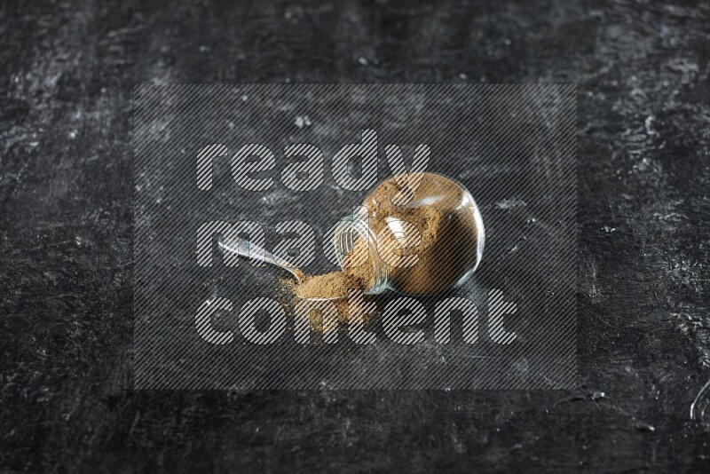 A flipped glass spice jar and a metal spoon full of cumin powder and powder spilled out on a textured black flooring