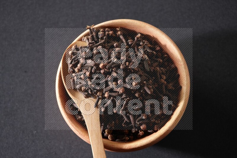 A wooden bowl and a wooden spoon full of cloves on a black flooring