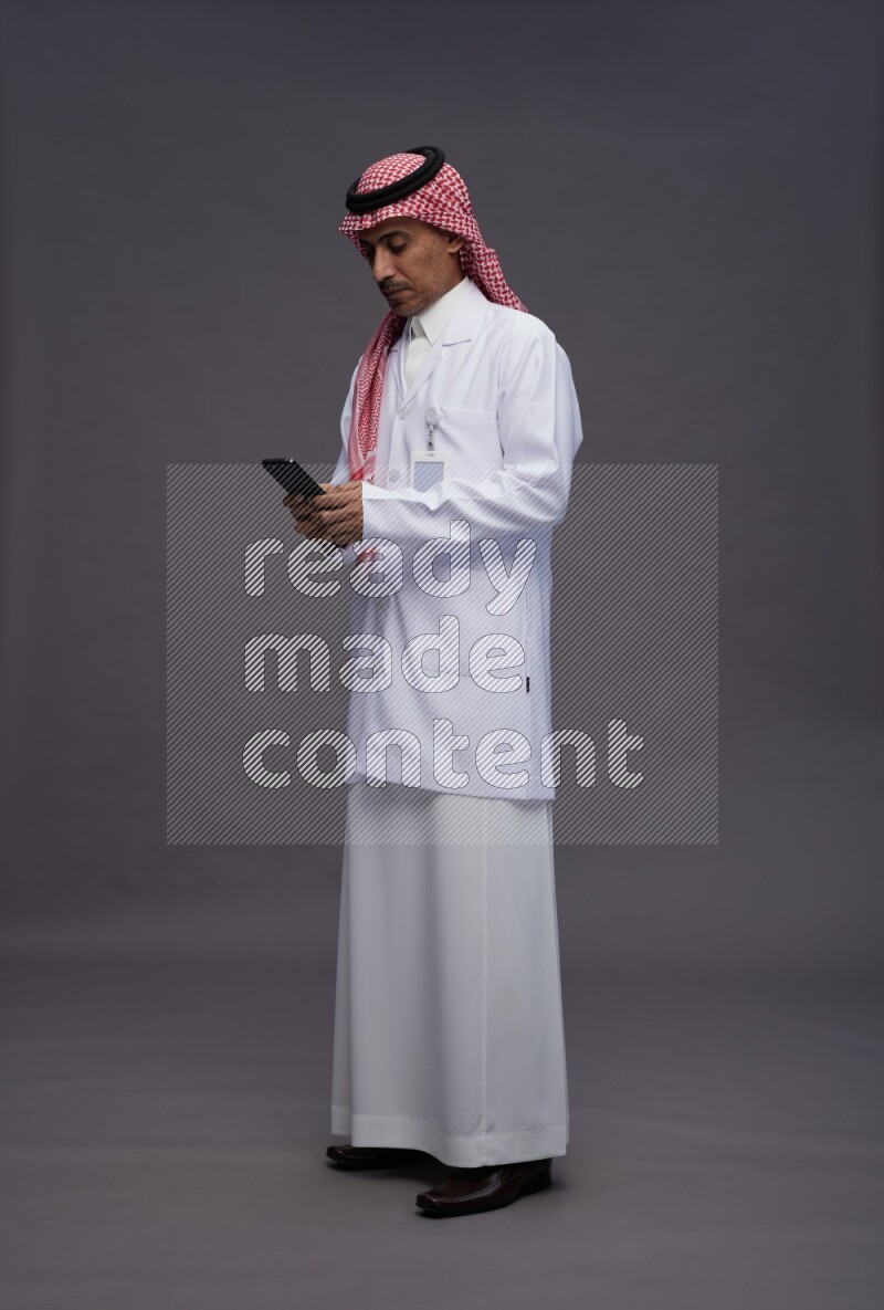 Saudi man wearing thob with lab coat and shomag with pocket employee badge standing texting on phone on gray background