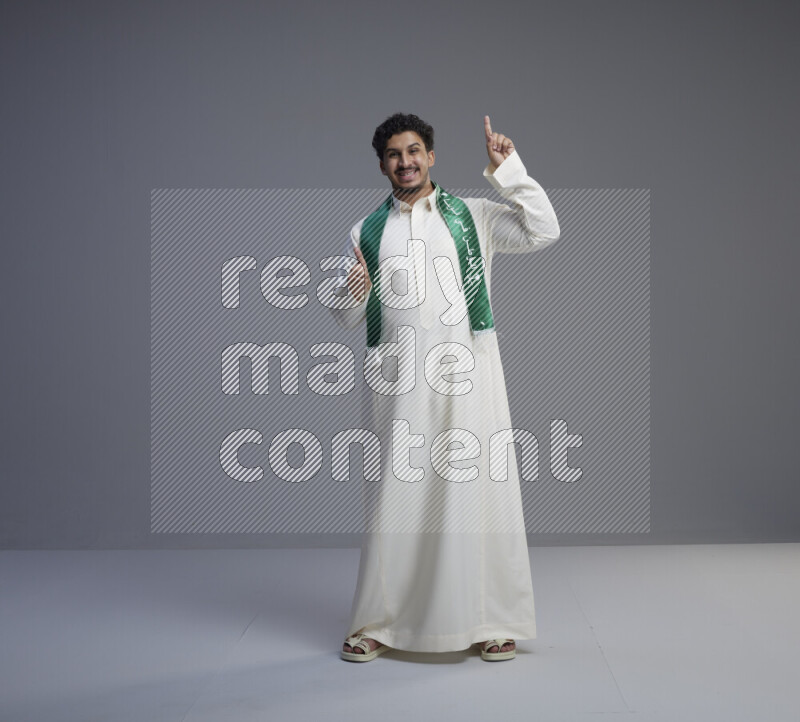 A Saudi man standing wearing thob and Saudi flag scarf on gray background