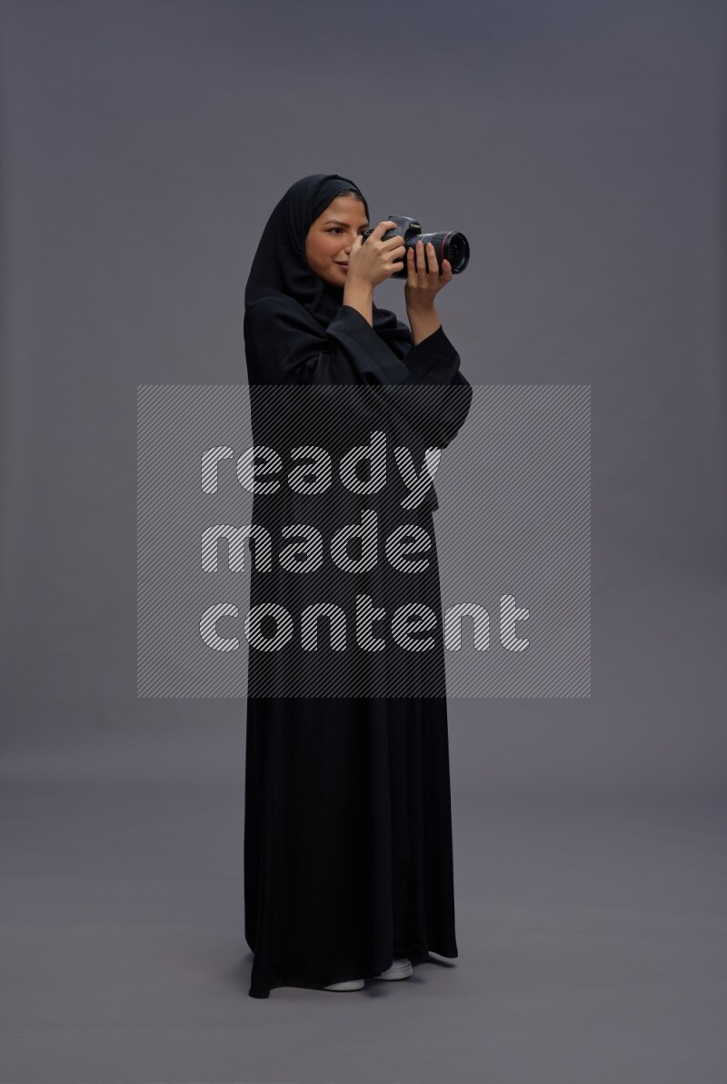 Saudi woman wearing Abaya standing holding Camera on gray background