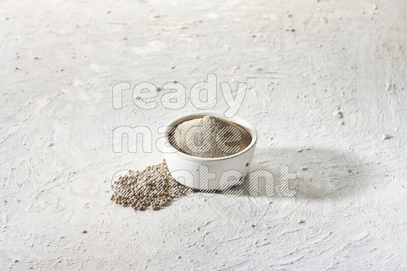 white pottery bowl full of white pepper powder with pepper beads on textured white flooring