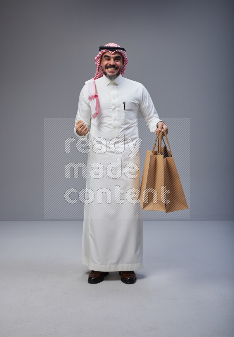 Saudi man Wearing Thob and red Shomag standing holding shopping bag on Gray background