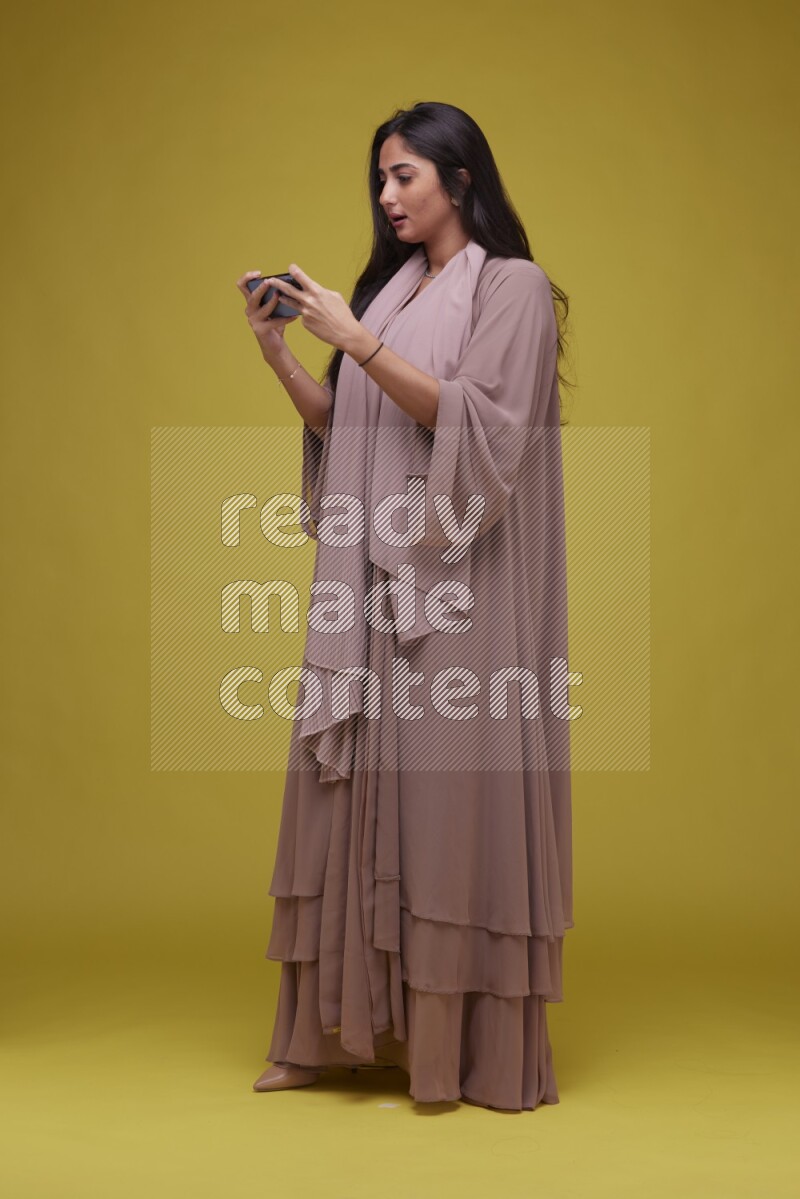 A woman Playing a Game on her smartphone on a Yellow Background wearing Brown Abaya