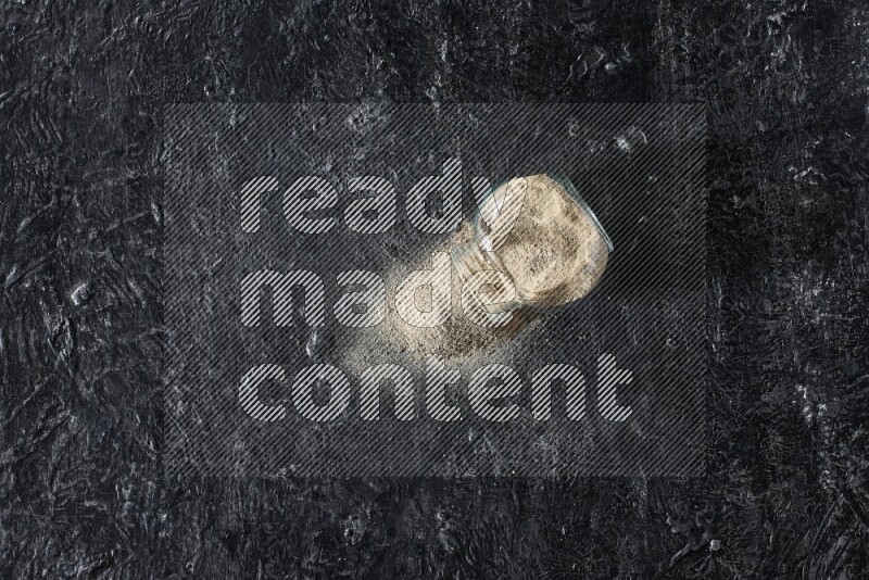 A flipped herbal glass jar full of white pepper powder with spilled powder on textured black flooring