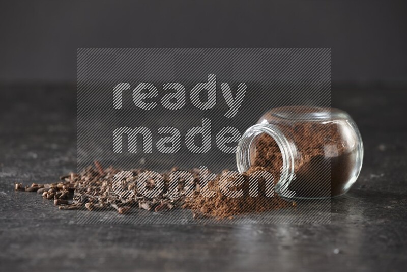 A flipped glass spice jar full of cloves powder and powder came out of it with cloves spread on textured black flooring