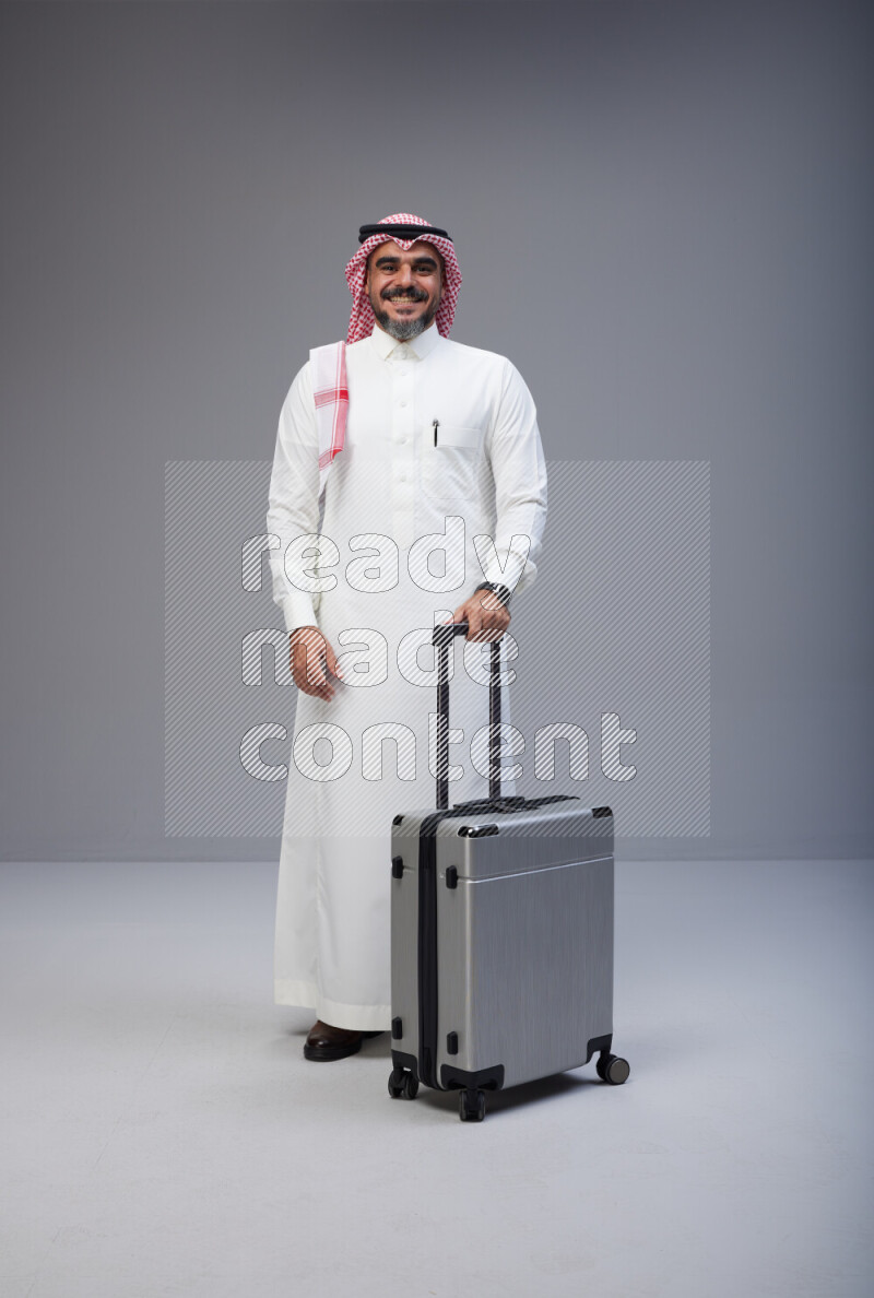 Saudi man wearing Thob and red Shomag standing holding Travel bag on Gray background