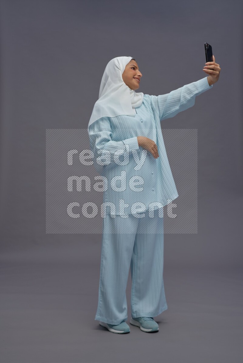 Saudi woman wearing hijab clothes standing taking selfie on gray background