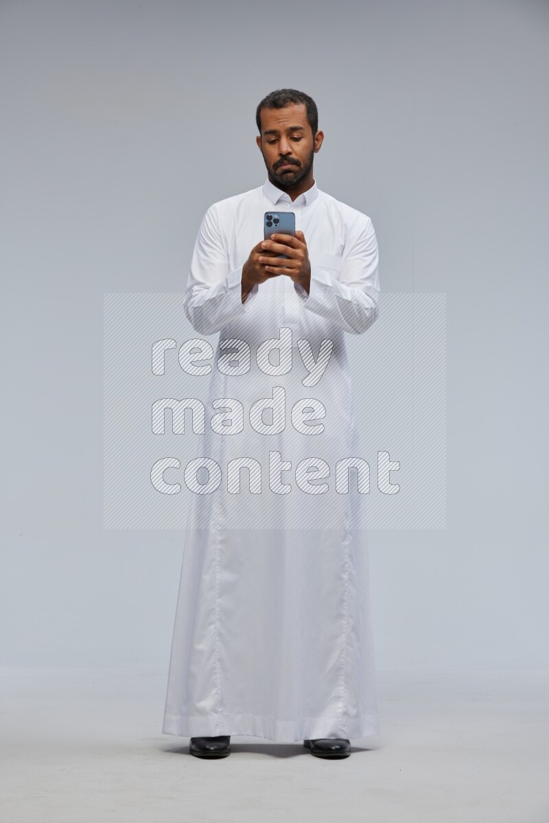 Saudi man wearing Thob standing texting on phone on Gray background