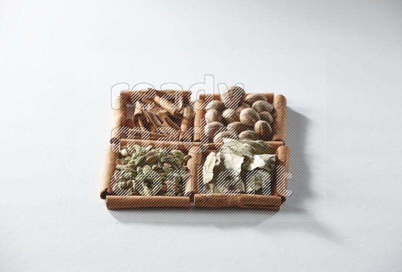 4 مربعات من أعواد القرفة ممتلئة بجوزة الطيب وأوراق الغار وحبوب الهيل والقرفة على أرضية بيضاء