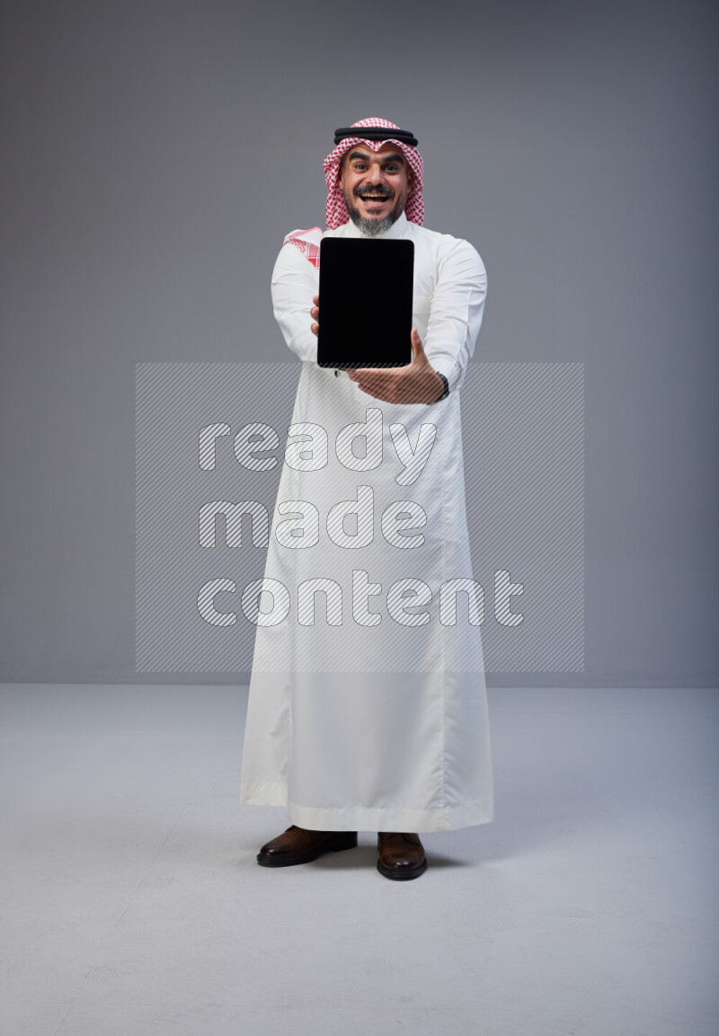 Saudi man Wearing Thob and red Shomag standing showing tablet to camera on Gray background