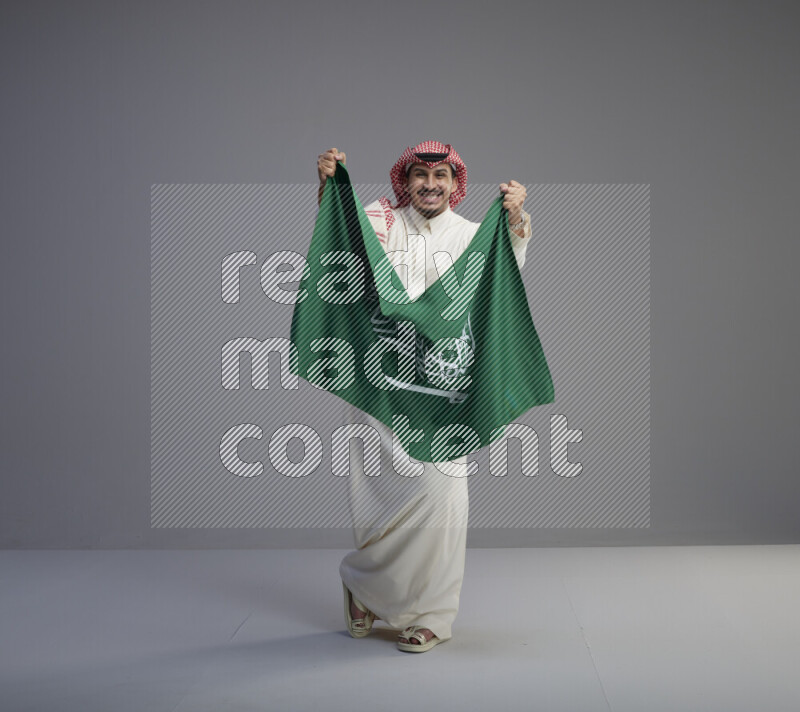 A saudi man standing wearing thob and red shomag holding big saudi flag on gray background