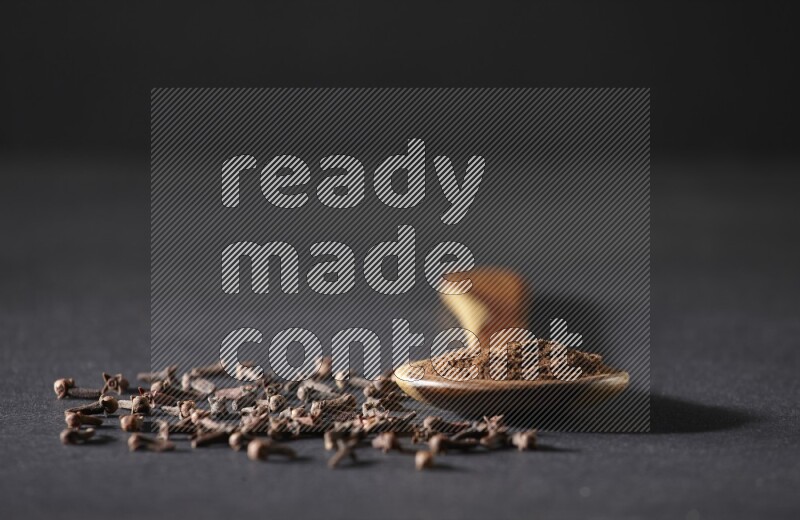 A wooden ladle full of cloves powder and some of whole cloves around it on a black flooring