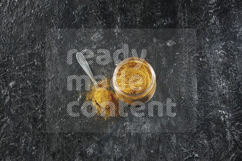 A glass jar and a metal spoon full of turmeric powder on a textured black flooring