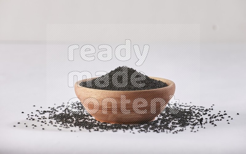 A wooden bowl full of black seeds and more seeds spread on a white flooring
