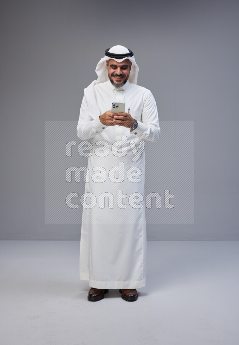 Saudi man Wearing Thob and white Shomag standing texting on phone on Gray background