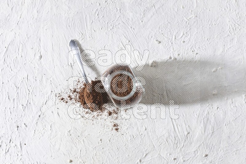 A glass spice jar and a metal spoon full of cloves powder on textured white flooring