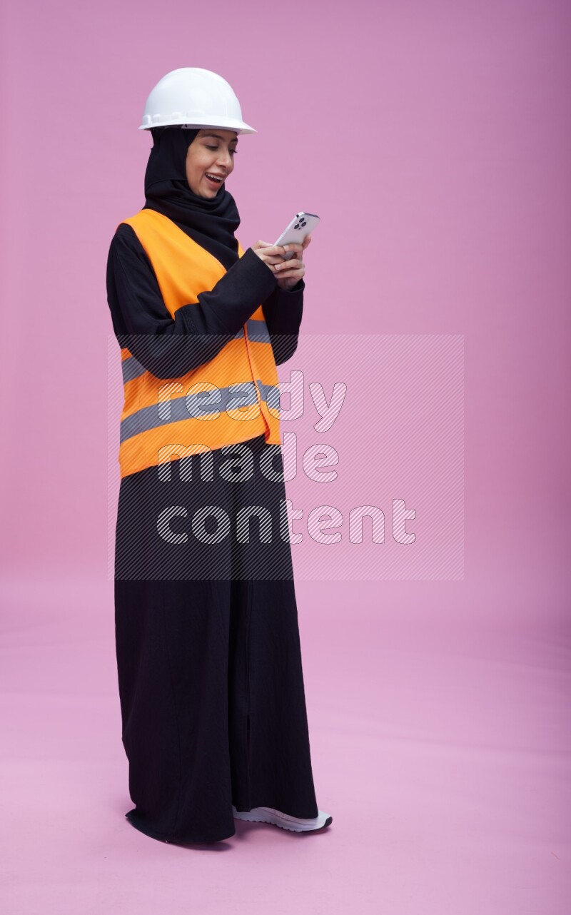 Saudi woman wearing Abaya with engineer vest and helmet standing texting on phone on pink background
