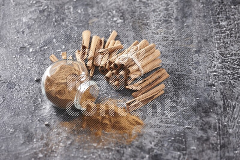 Herbal glass jar full of cinnamon powder flipped with cinnamon sticks stacked and bounded on a textured black background