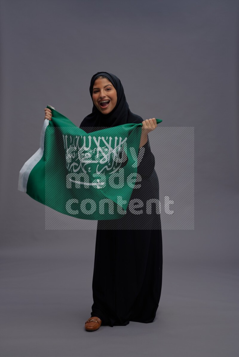 Saudi woman wearing Abaya standing holding Saudi flag on gray background