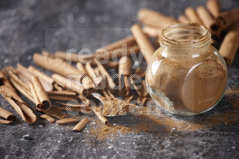 Herbal glass jar and a metal spoon full of cinnamon powder surrounded by cinnamon sticks on textured black background