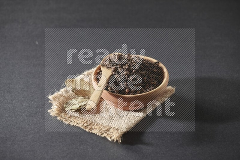 A wooden bowl, a wooden spoon full of cloves, and bay leaves (laurel) on a piece of burlap on a black flooring