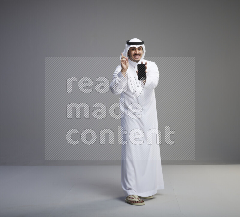 A Saudi man standing wearing thob and white shomag showing phone to camera on gray background