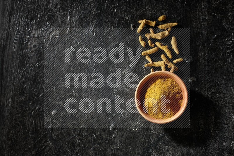 A wooden bowl full of turmeric powder with dried turmeric fingers on textured black flooring