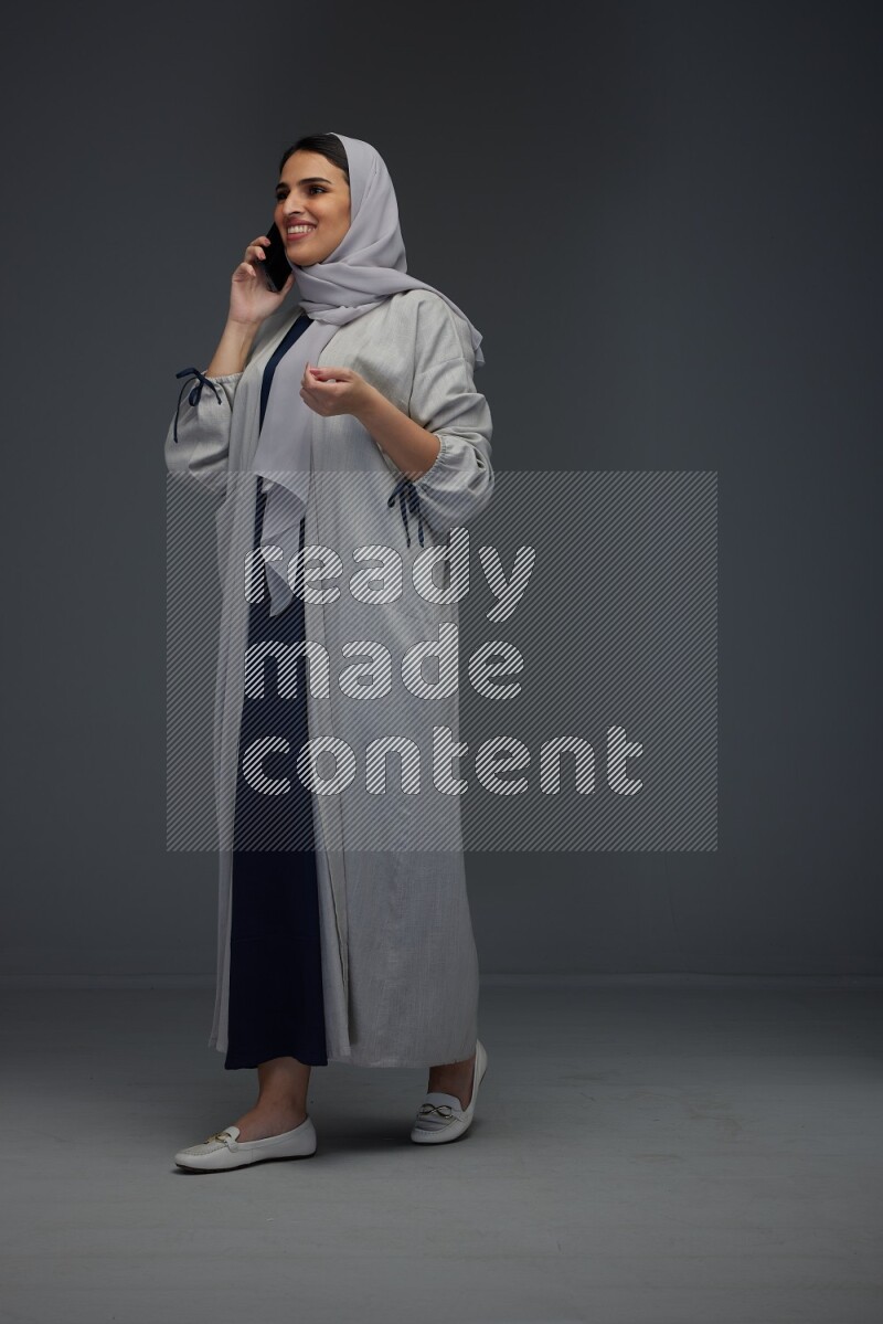 A Saudi woman wearing a light gray Abaya and head scarf standing and talking in the phone eye level on a grey background