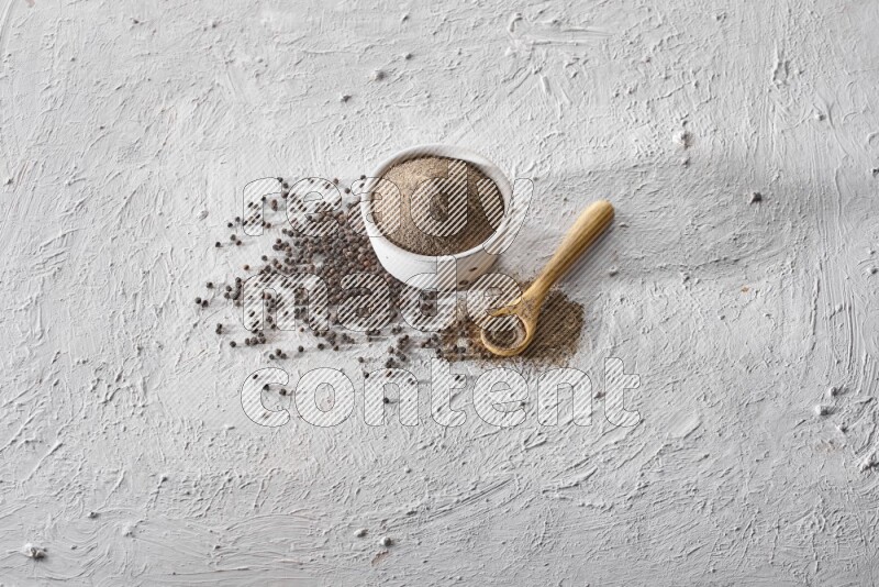A pottery white bowl full of black pepper powder and wooden spoon full of powder and black pepper beads on a textured white flooring