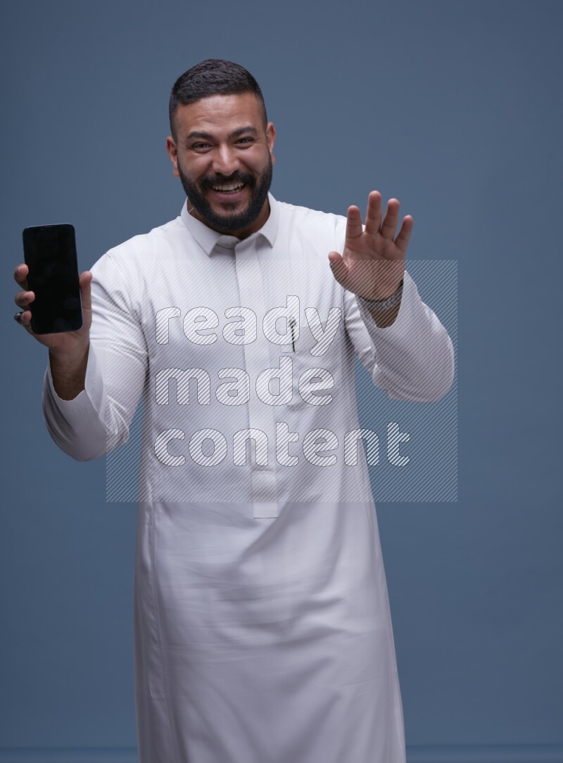 A man Showing his phone in Blue Background wearing Saudi Thob