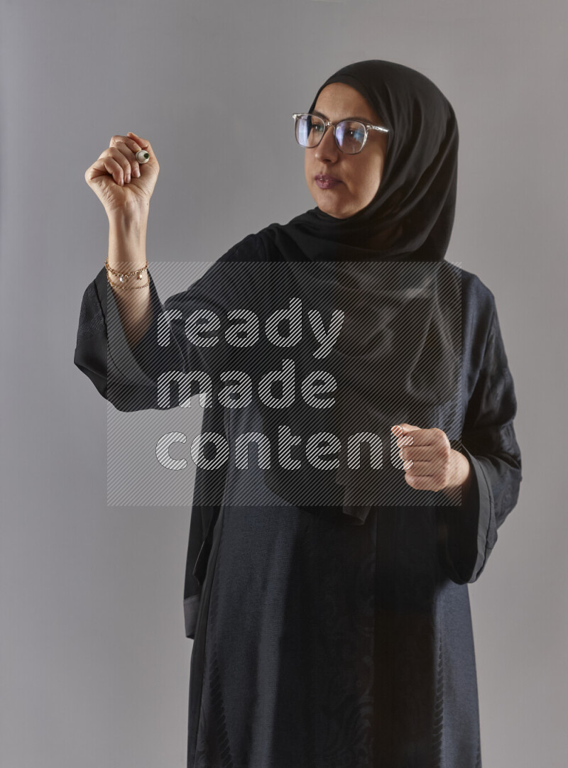 Woman in black abaya, hijab and glasses holding a marker pen to write on a transparent board with different reactions and expressions on grey background