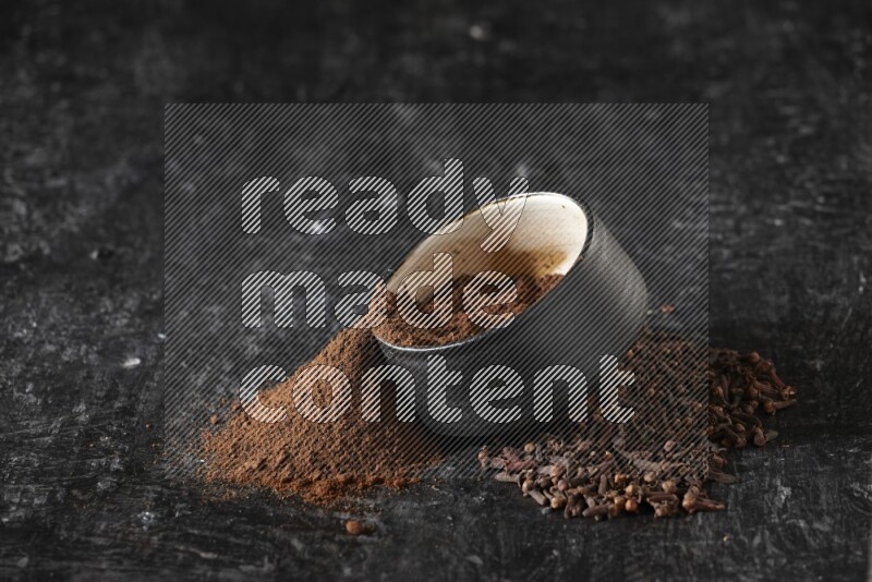 A black pottery bowl full of cloves powder spilled out of it with whole grains on a textured black flooring