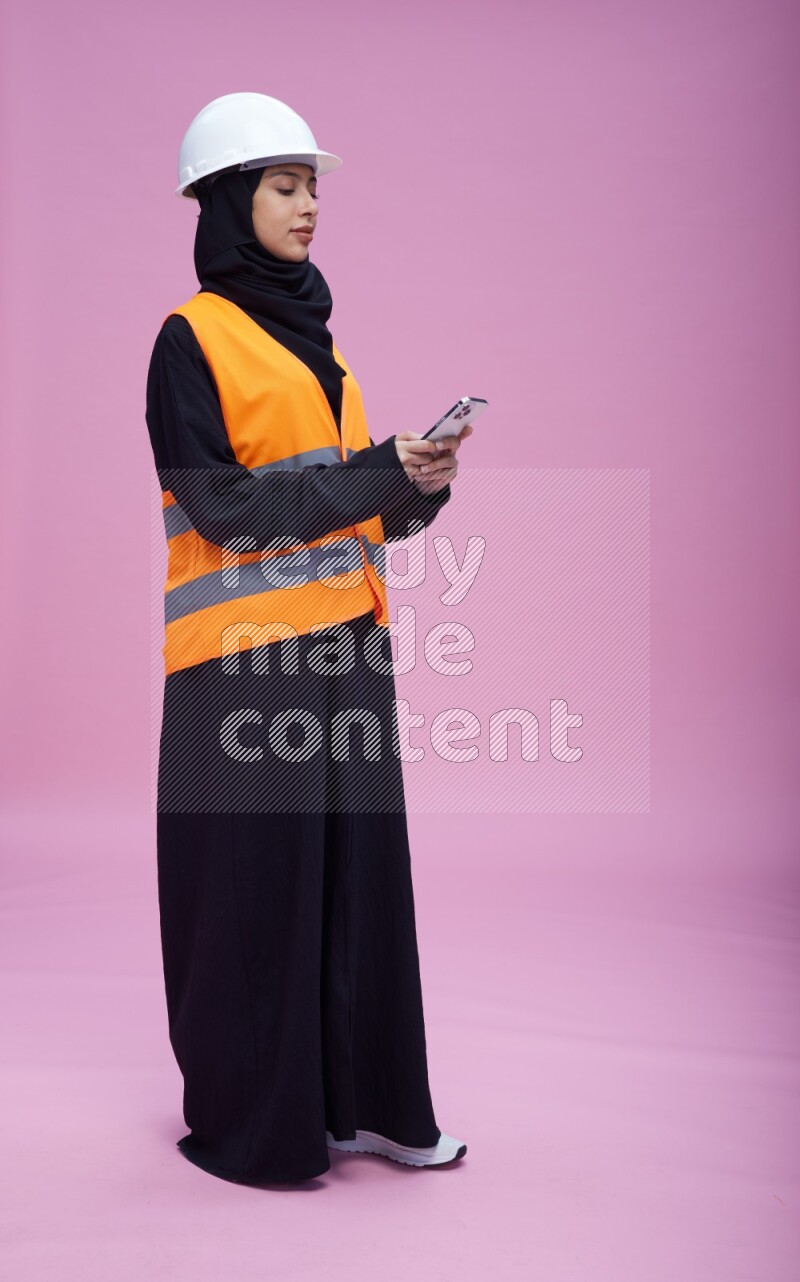 Saudi woman wearing Abaya with engineer vest and helmet standing texting on phone on pink background